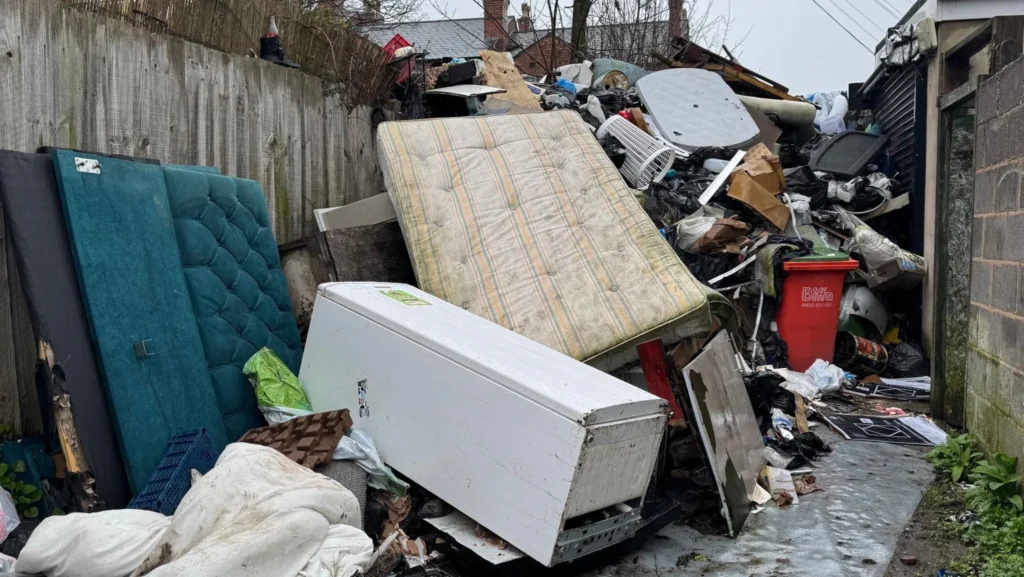 BBC An alleyway between a concrete wall and garden fences. The alleyway is full of dumped rubbish including mattresses, fridges, sofas, duvets and other household waste