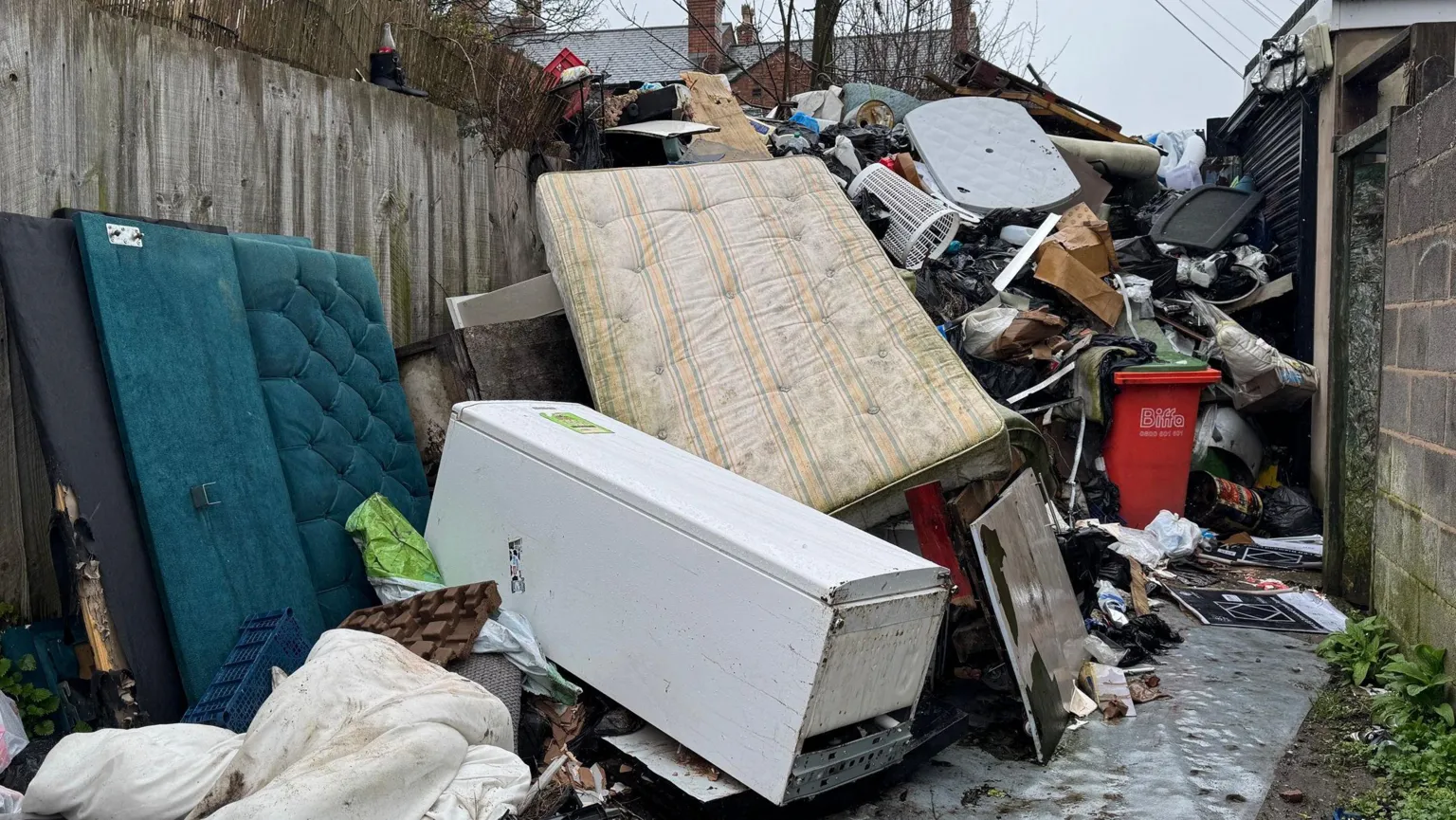 BBC An alleyway between a concrete wall and garden fences. The alleyway is full of dumped rubbish including mattresses, fridges, sofas, duvets and other household waste