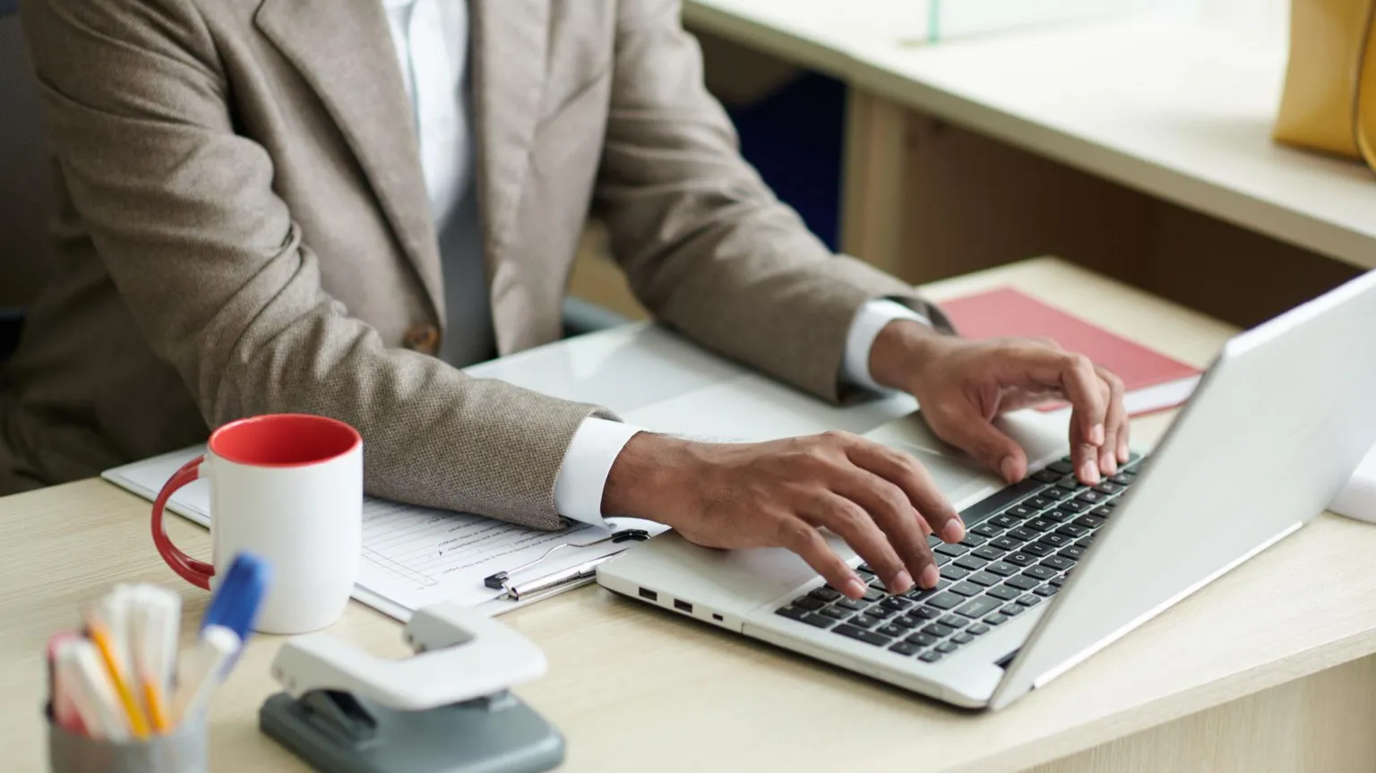 Getty Images A close up of a business person in a brown suit and white shirt (we only see from the neck down) typing on a grey laptop on a desk. The desk has a white and red mug, a hole puncher and stationary.
