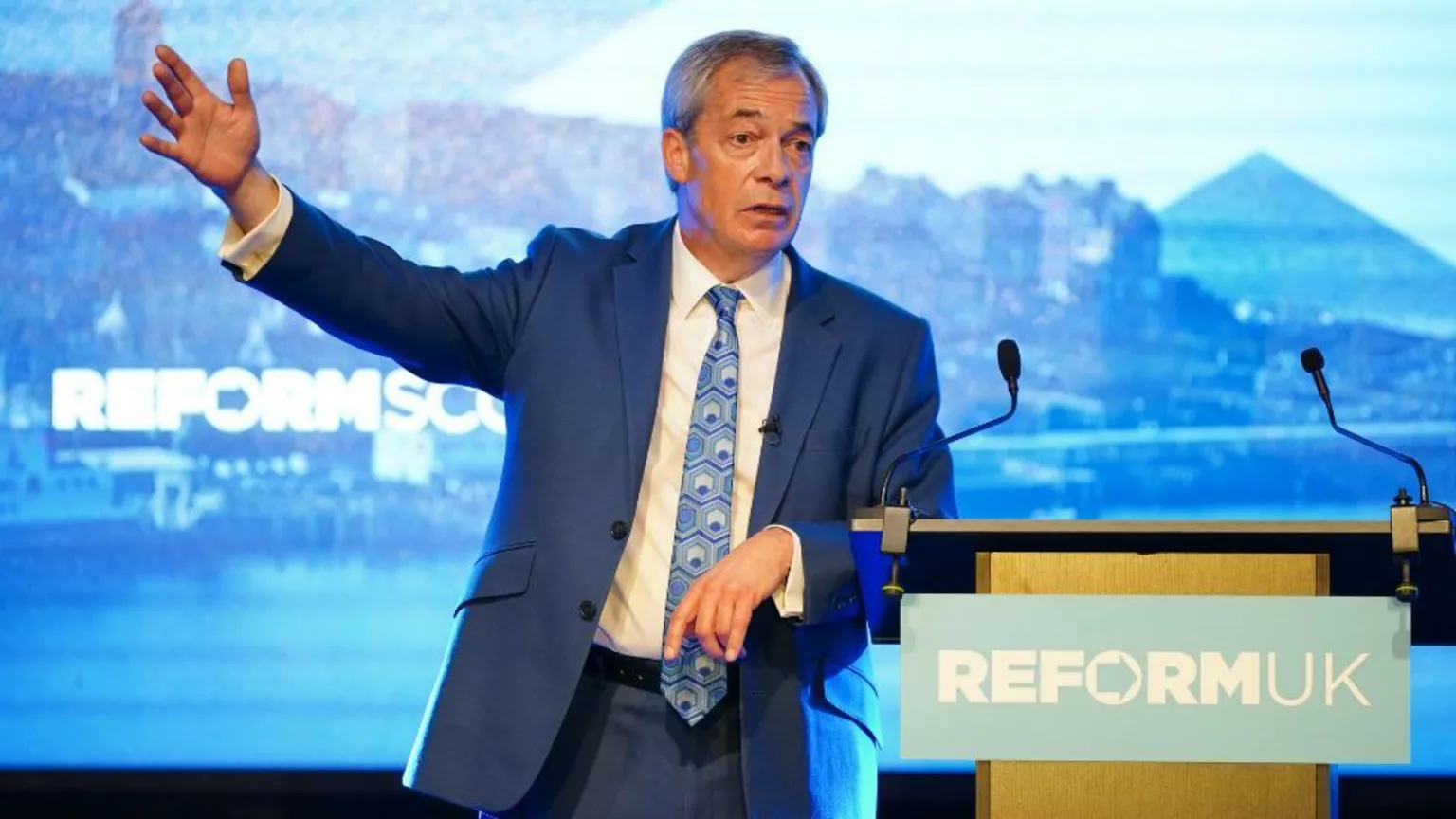 PA Media Nigel Farage, who has thinning grey hair, leans on a podium with his right arm outstretched. He is in front of a blue screen.