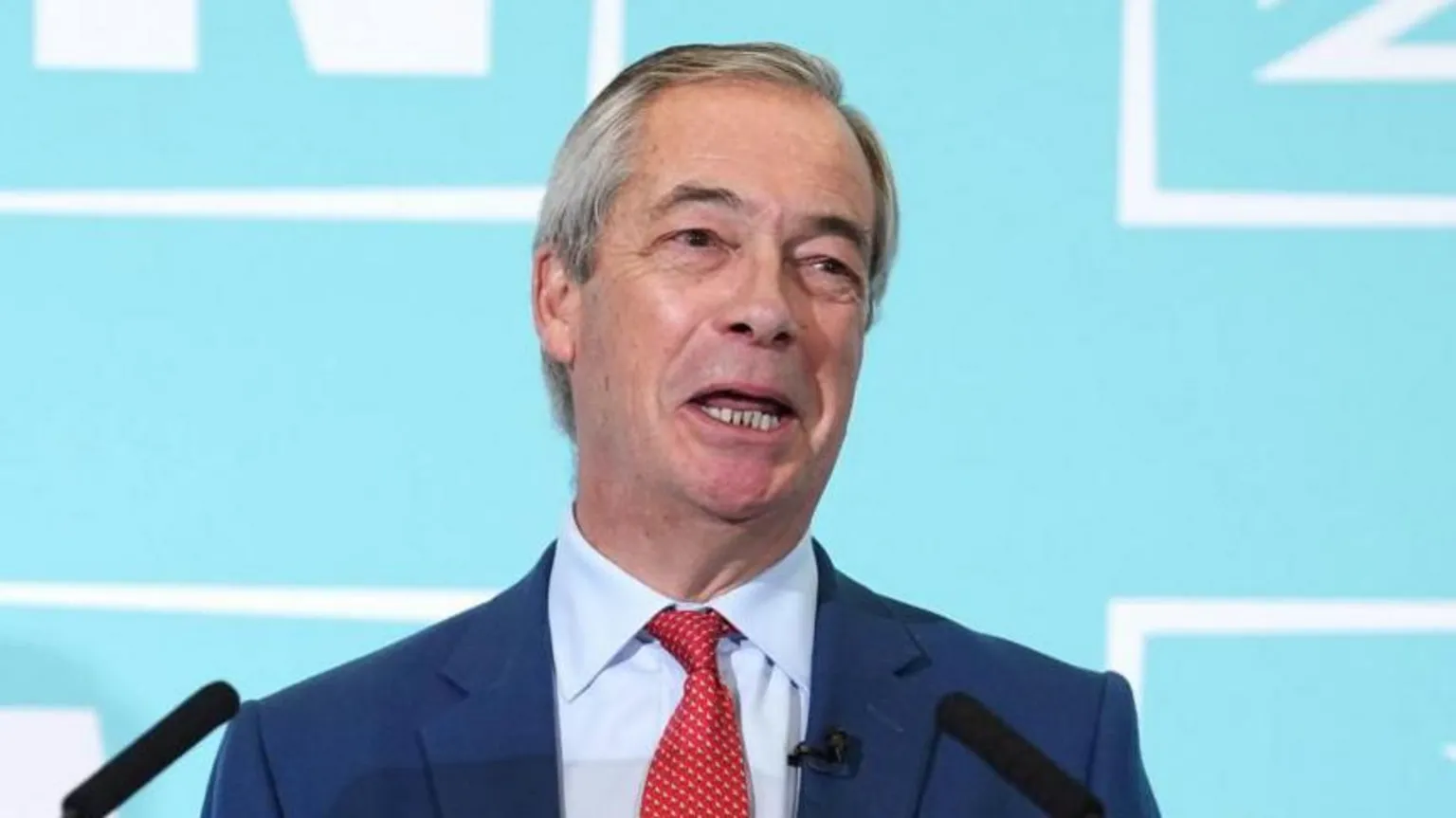 PA Reform Party leader Nigel Farage speaking during a Reform UK news conference. He is smiling and wearing a dark blue suit jacket, light blue shirt and red tie.
