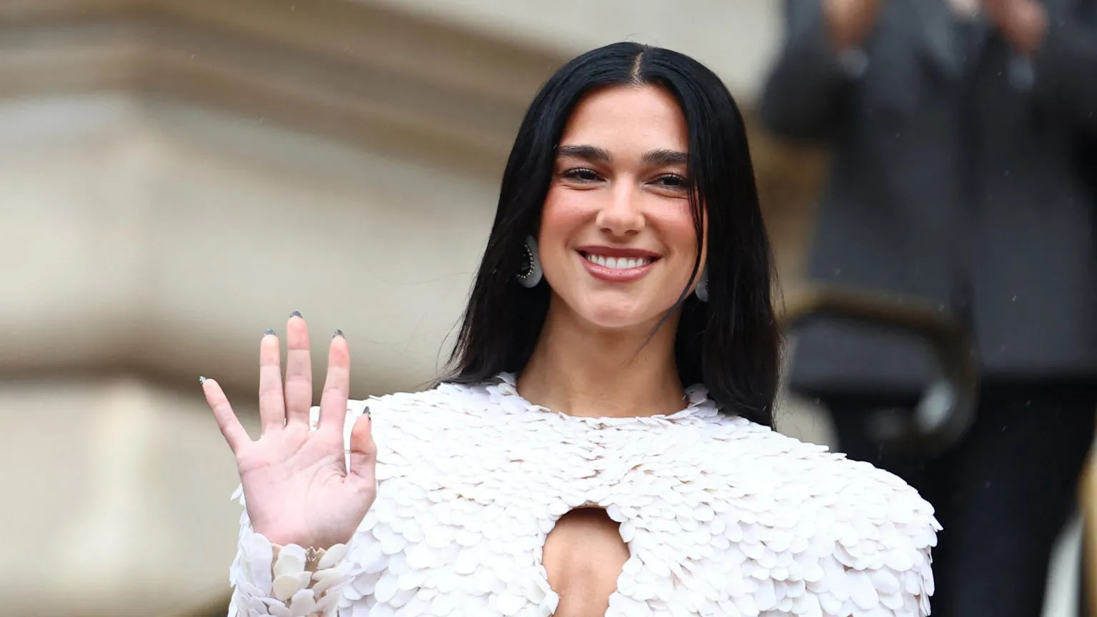 Reuters Dua Lipa wearing a feathered white outfit, waving at photographers and smiling.