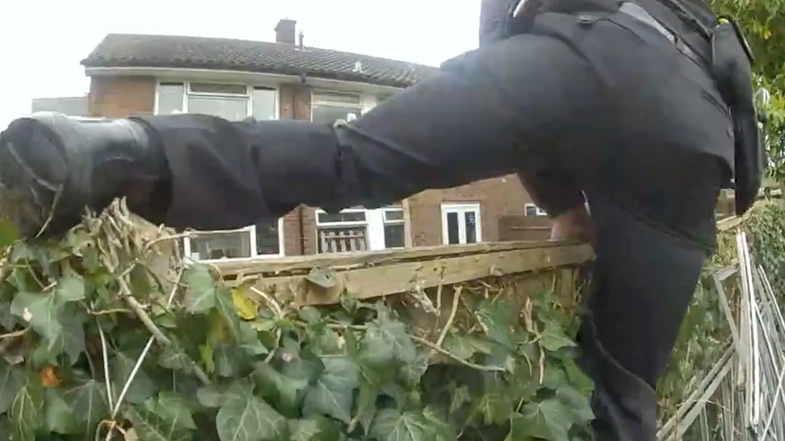 Still image of a police officer, dressed all in black, as he climbs over a wooden fence. The fence is covered in green ivy.
