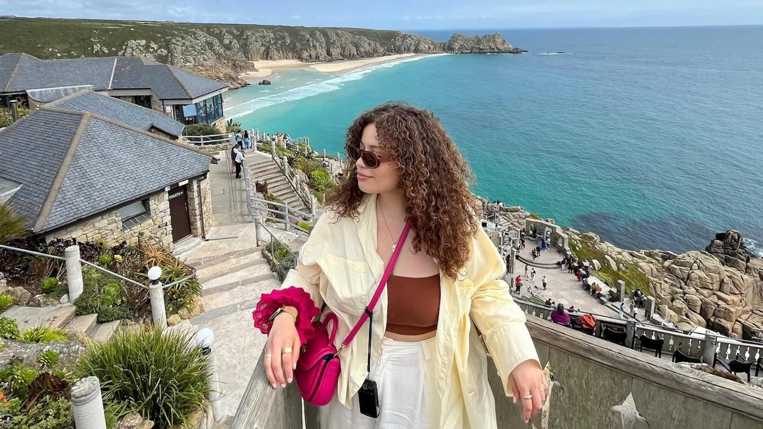@eboniivoryblog Eboni a woman in her 30s with curly brown hair standing above The Minack Theatre which is carved into the cliff face in Cornwall and covered in luscious green gardens with a view across turquoise sea