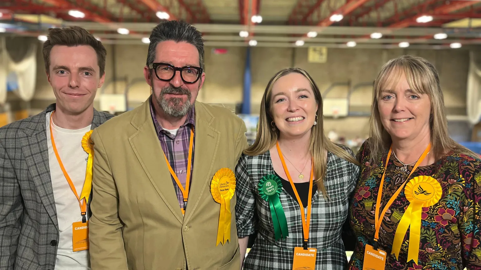 A son, father, daughter and mother smile at the camera. All are wearing orange lanyards which say "candidate". The daughter wears a Green party rosette, whilst the other three wear Liberal Democrat rosettes. The photo has been taken in a sports hall during an election count.