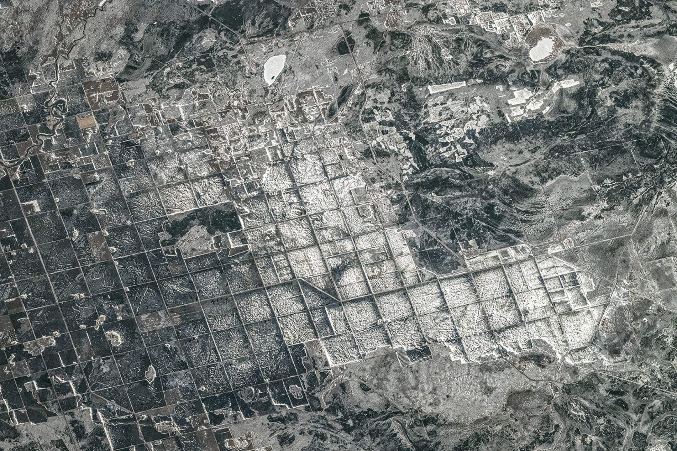 A satellite image shows a mostly grayscale landscape featuring grid-like patterns contrasted against surrounding irregular terrain.