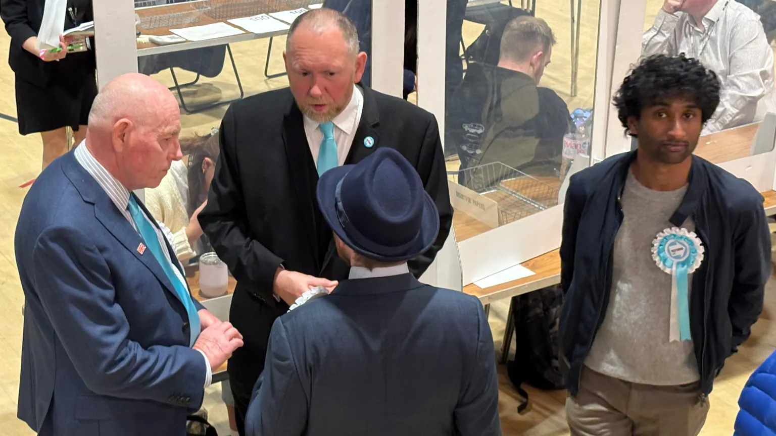 Four men, one in a blue suit, one in a black suit, one in a dark jacket and a trilby hat, and one with a dark casual jacket over a grey T-shirt, are talking in a huddle. Reform UK rosettes are visible on three of them. One has dark hair and a beard and is looking at the camera.