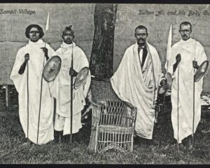 A black-and-white photo of Somali warriors posing next to a wicker chair