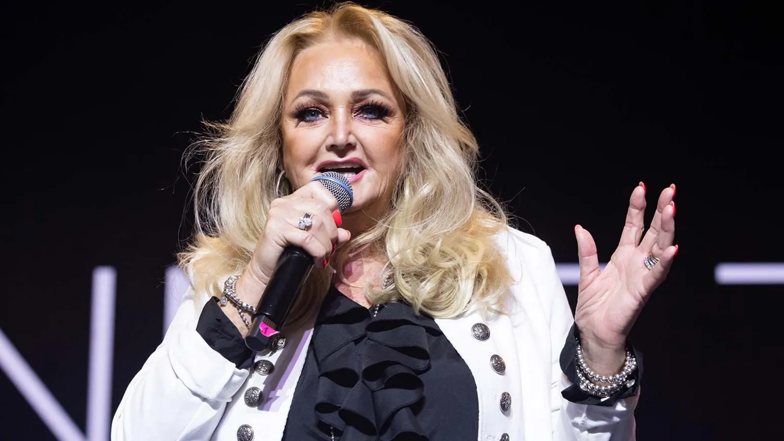 Bonnie Tyler on stage holding a microphone. She has long blonde hair, and is wearing a black blouse and a white  jacket