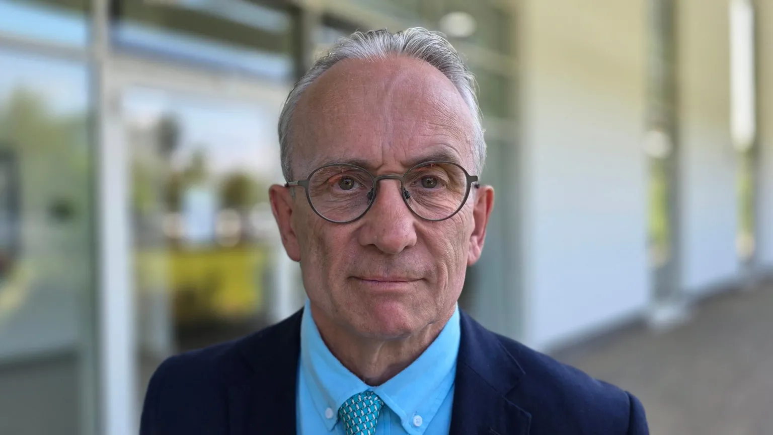 David Bick is wearing a dark blue suit jacket on top of a lighter blue shirt. He also has a blue patterned tie.