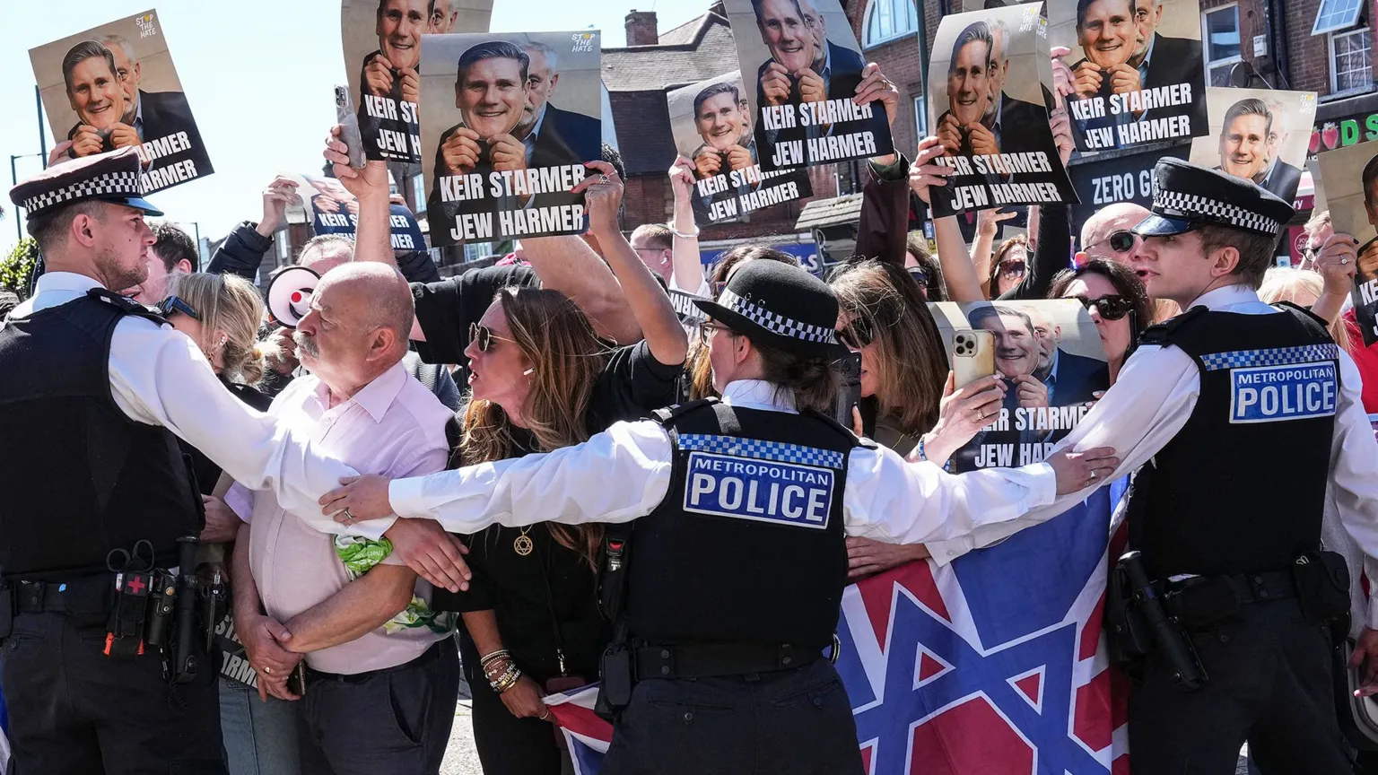 Protesters holding up placards with Keir Starmer's face  printed on them being held back by police in  Golders Green today 3oth April 2026