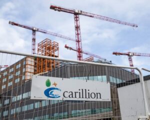 A Carillion sign on a fence in front of a construction site with red tower cranes and scaffolding above