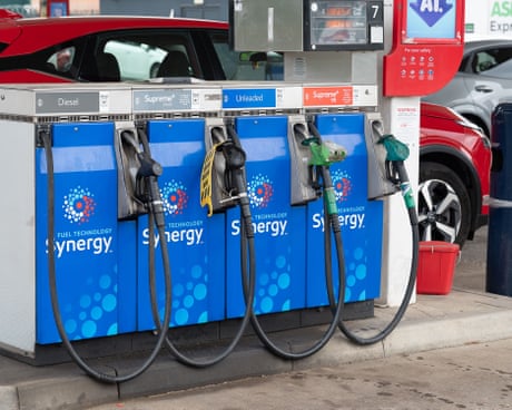 Four blue-branded fuel pumps with 'Synergy' branding at a petrol station, with cars visible in the background.