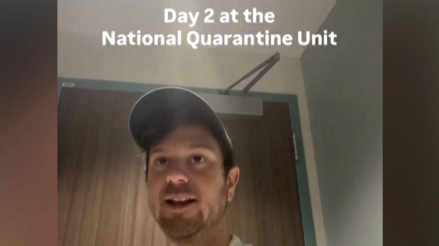 A man in a baseball cap below on-screen text that reads "Day 2 at the National Quarantine Unit".