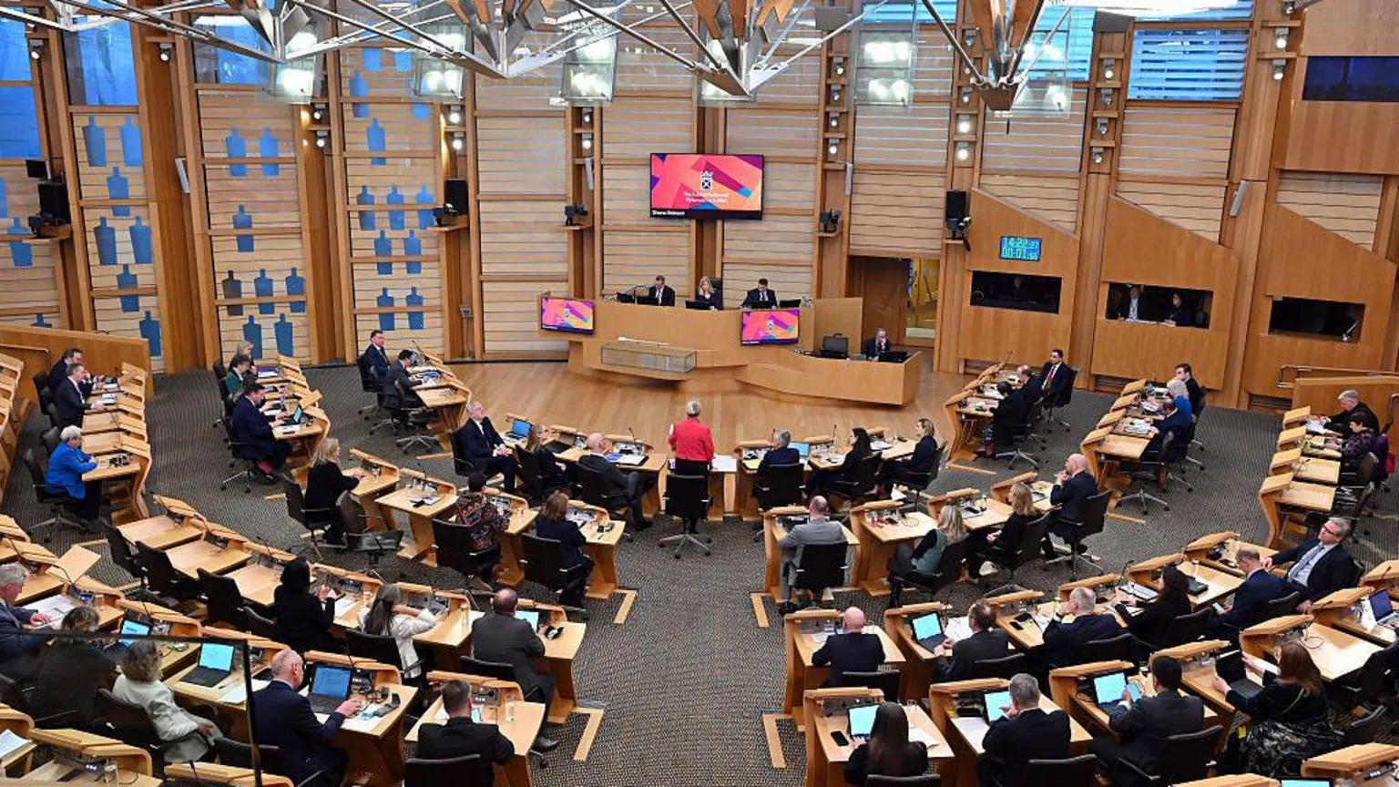 Getty Images Finance Secretary Shona Robison (standing in the centre wearing a red jacket) delivers the Scottish Budget for 2026-2027 in the chamber of the Scottish Parliament in Edinburgh on 13 January, 2026. A number of MSPs can be seen at wooden desks with computer screens.