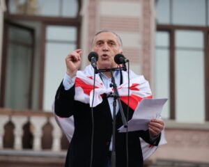 Paata Burchuladze speaks into a pair of microphones outside a large building with a Georgian flag tied round his shoulders
