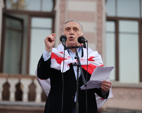 Paata Burchuladze speaks into a pair of microphones outside a large building with a Georgian flag tied round his shoulders