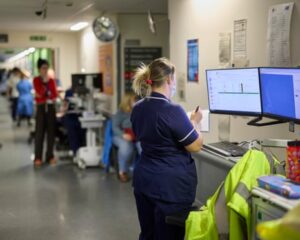 A nurse on an emergency ward wearing dark blue scrubs stands looking at computer screens and taking notes.