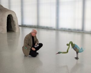 A man with thinning white hair kneels down on the floor of a gallery to look closely at a sculpture of a long-necked bird-like creature.
