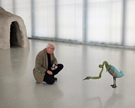 A man with thinning white hair kneels down on the floor of a gallery to look closely at a sculpture of a long-necked bird-like creature.