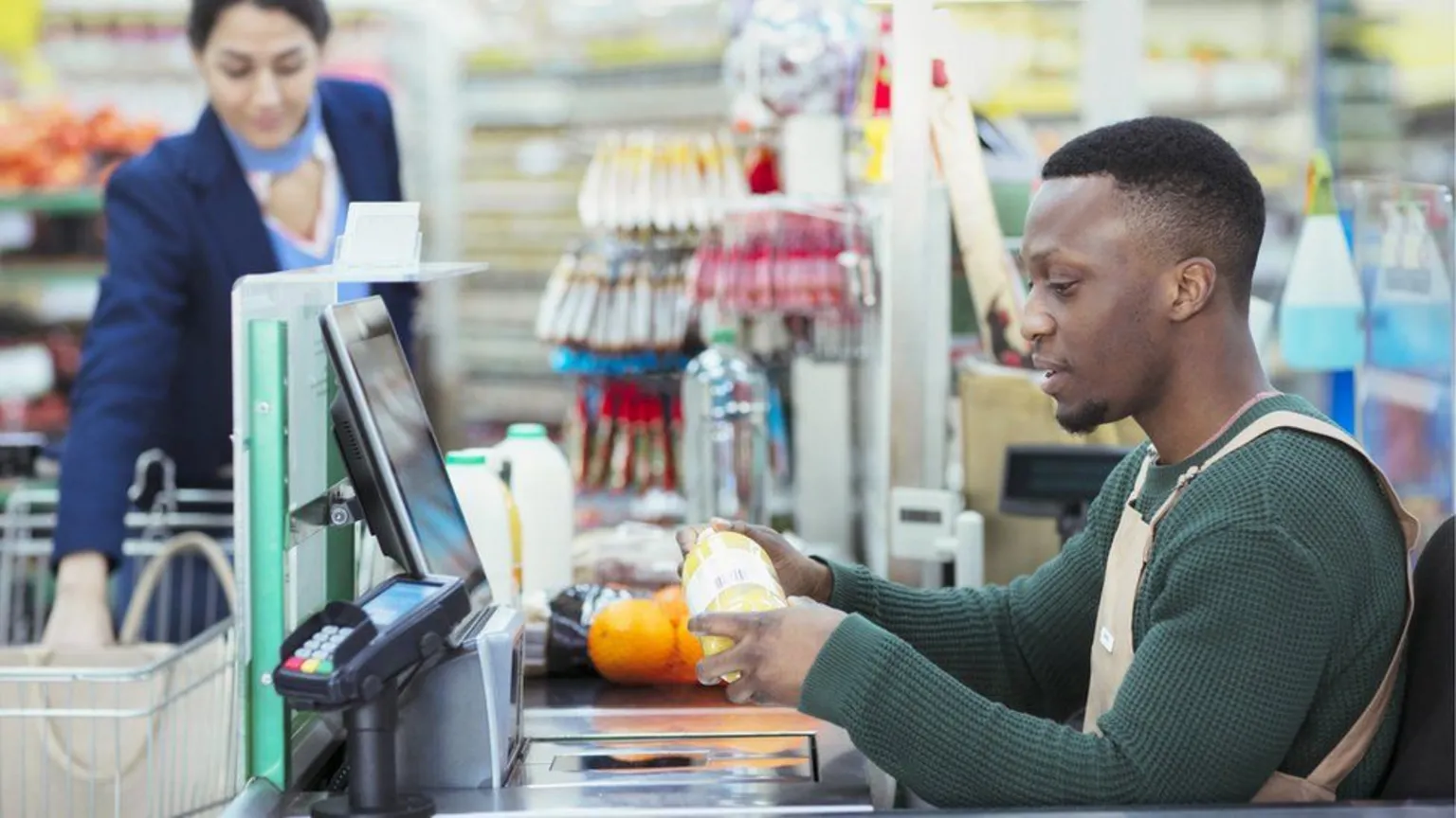 Getty Images A young man wearing a green sweater and a beige apron works on a supermarket checkout. He is serving a woman wearing a blue and pink top and navy jacket.