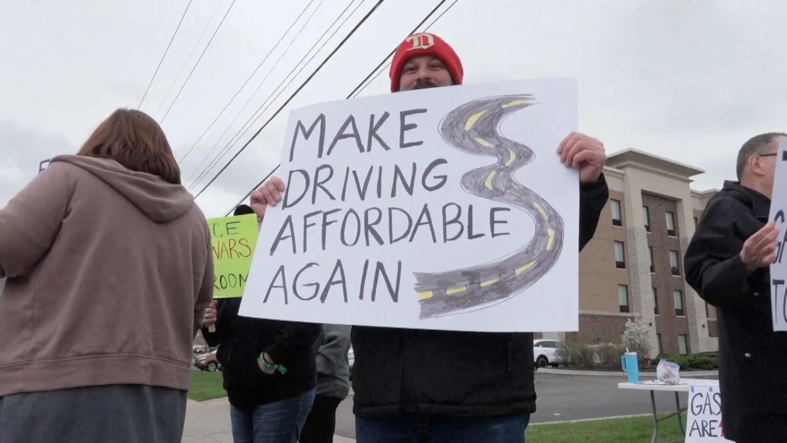 A man is holding up a sign that says "make driving affordable again".