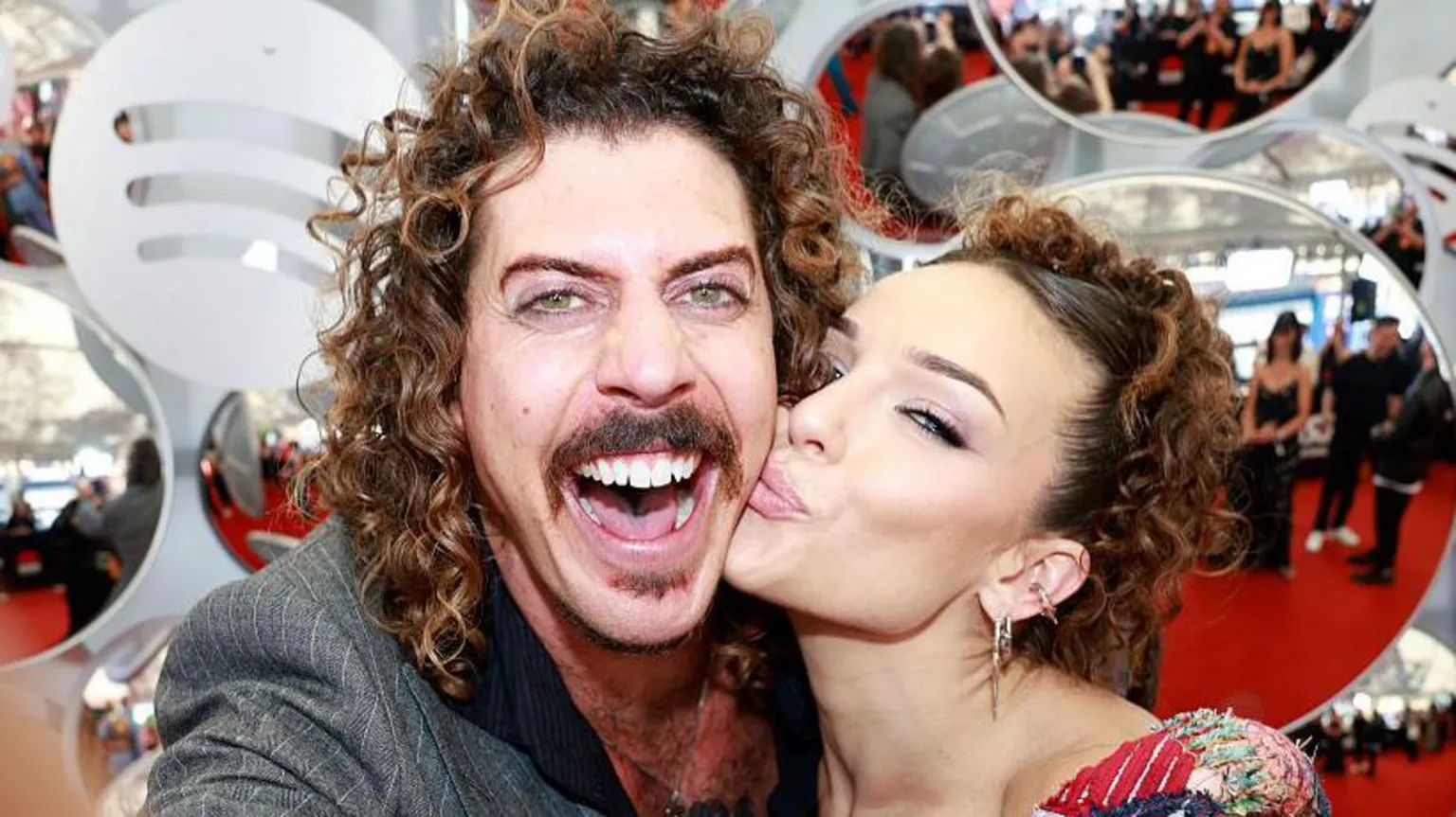 A man with curly brown hair, smiling widely at the camera, while a woman kisses him on the cheek