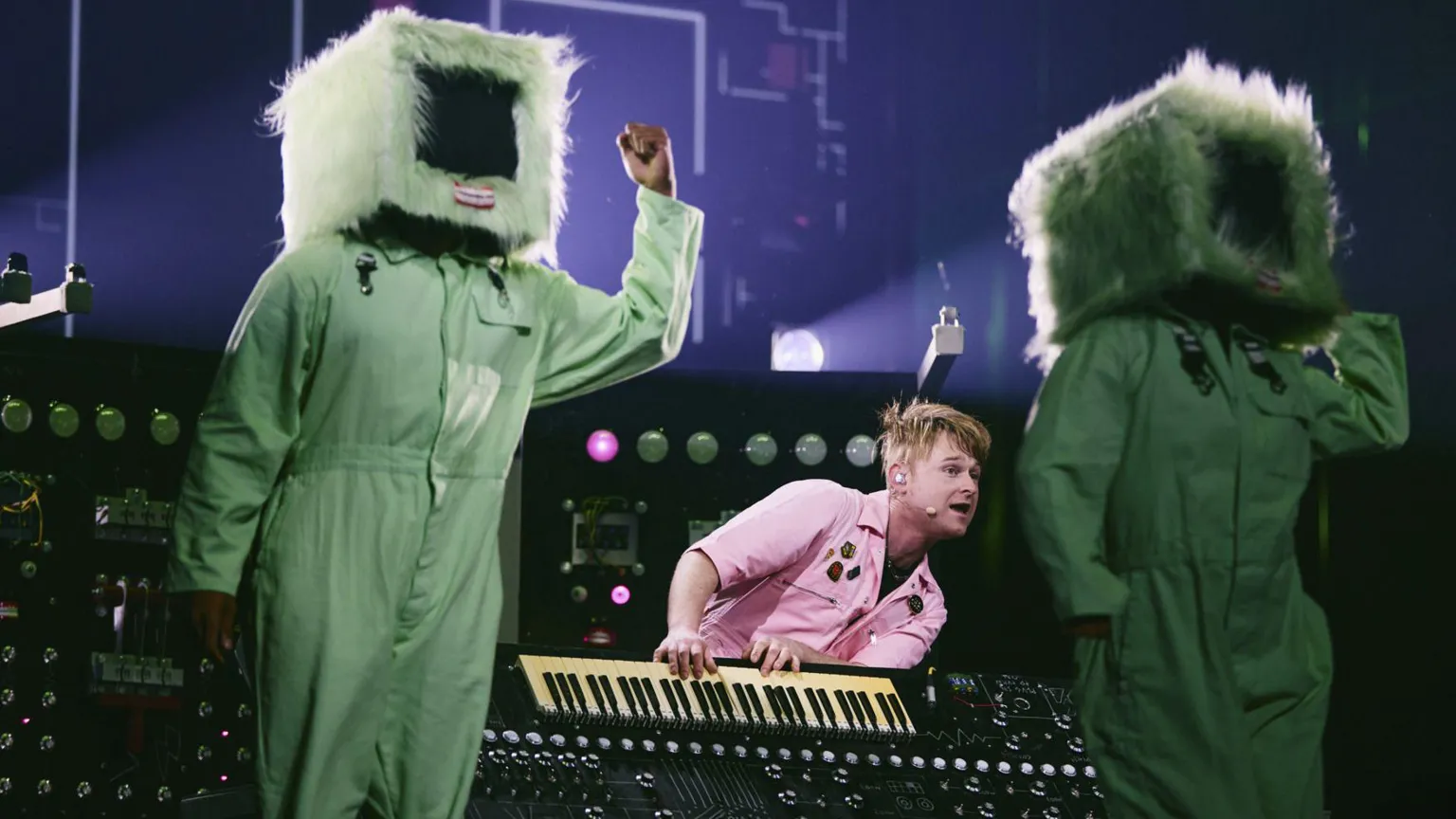 A man in a pink shirt playing a keyboard. On either side of him are two people, dressed in a green jumpsuit and a television prop over their heads.