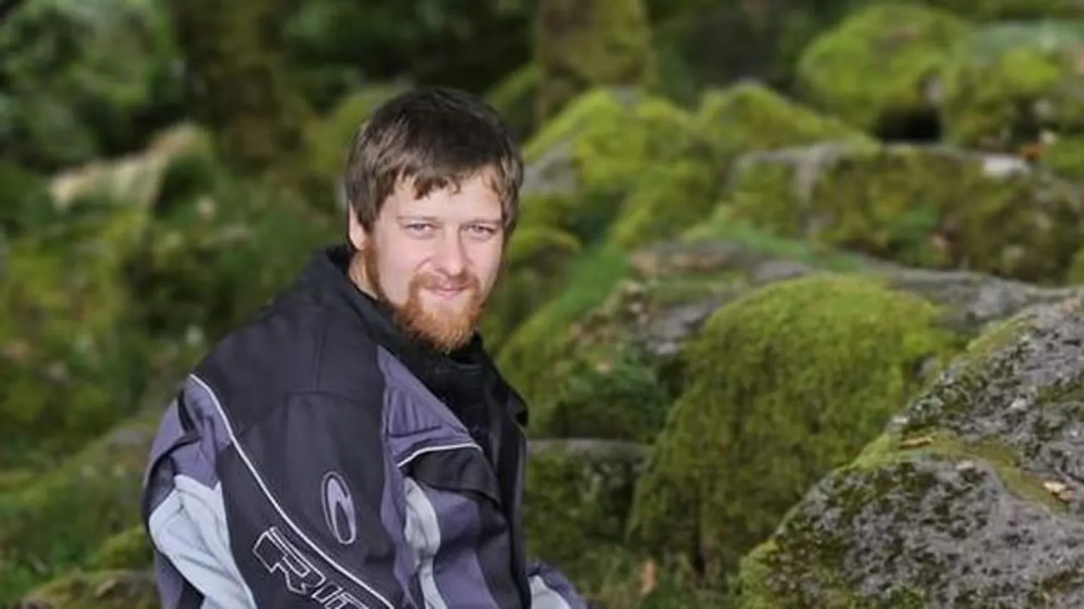 Ben Rowe, with a beard, is wearing a motorcycling jacket and sitting on rocks covered in moss in his former Facebook profile picture