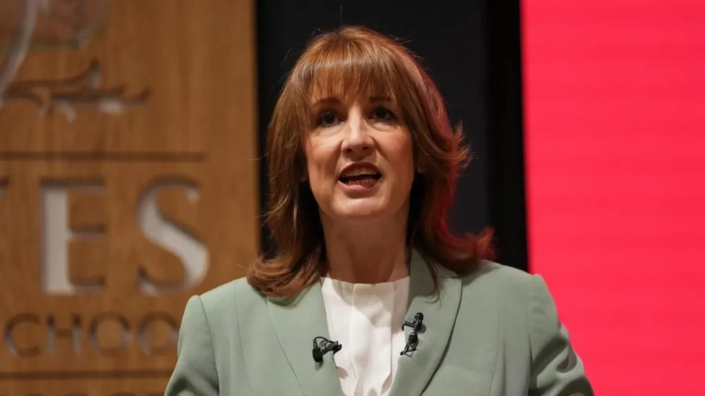 PA Media Rachel Reeves wears a sage green blazer while standing in front of a red-lit screen and a wooden sign.
