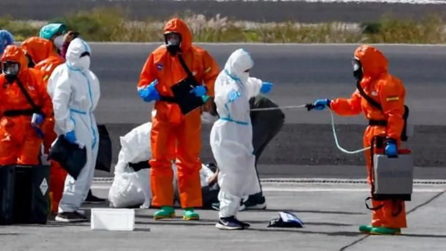People dressed in white and orange hazmat suits stand on the tarmac next to a plane in Tenerife. A person in an orange hazmat suit is hosing down another in a white suit, while others look on.