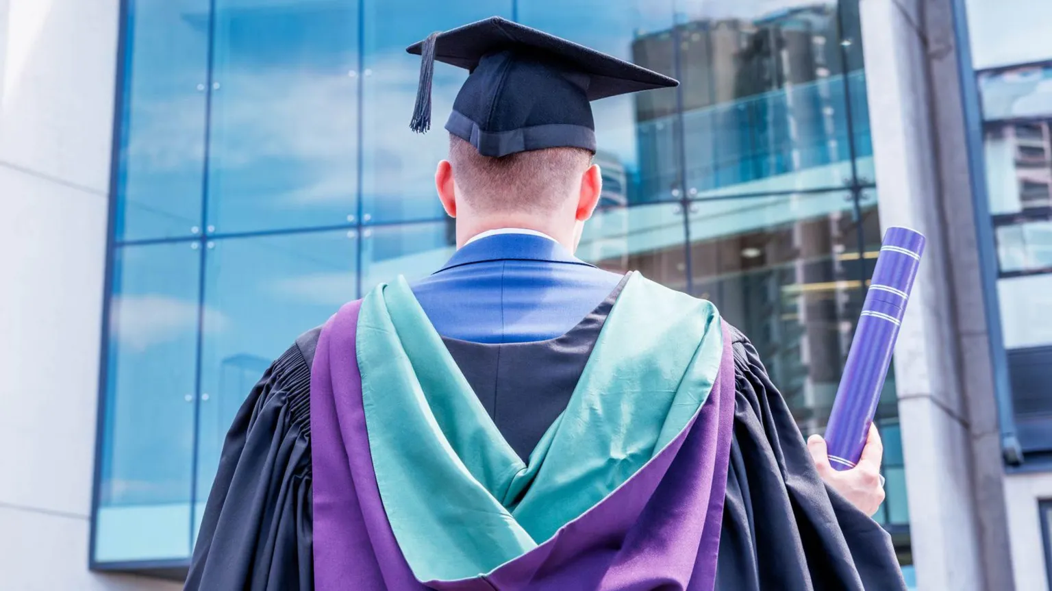 A graduate stands with his back to the camera. He is holding a cap and a scroll.