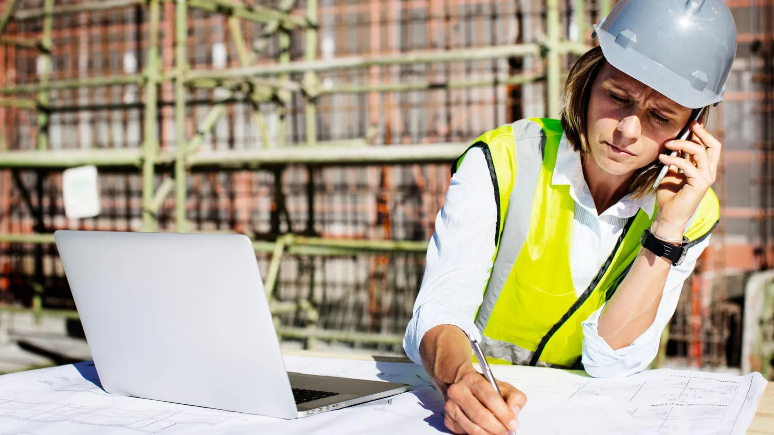 Getty Images A woman wearing a grey hard hat and hi vis writes on a plan while on the phone with her laptop open next to her with a buolding site in the background