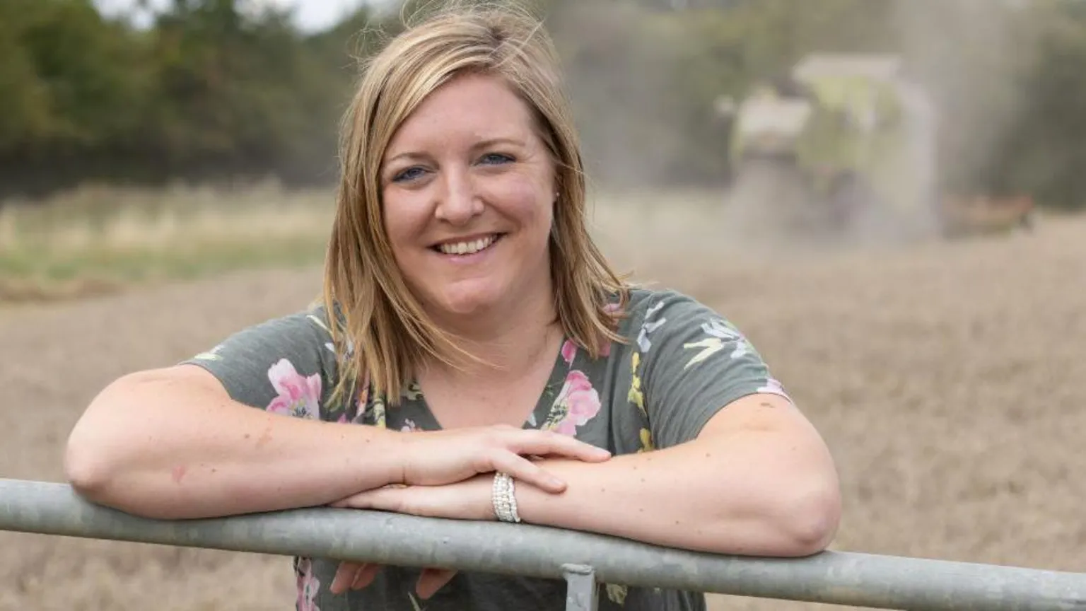 Milly leans on a metal farming gate and smiles at the camera. She has shoulder length blonde hair and wears a grey t-shirt with pink and yellow flowers. In the background, blurred, is a combine harvester and brown fiel.
