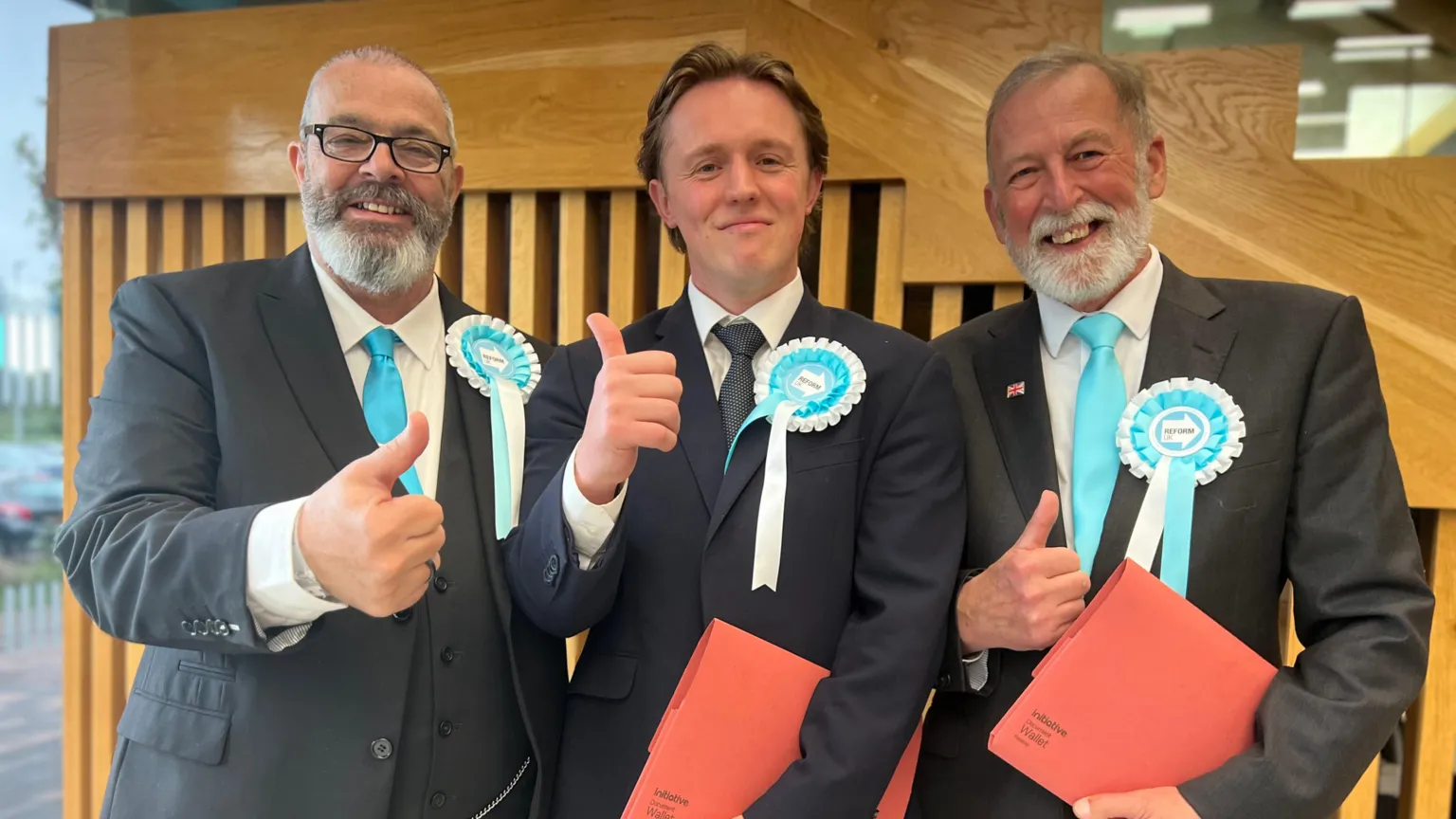 Three men in dark suits and light blue ties stand giving the thumbs up. They wear election rosettes identifying them as representing Reform UK and two of them hold folders. They are all white, two are older and one is younger.