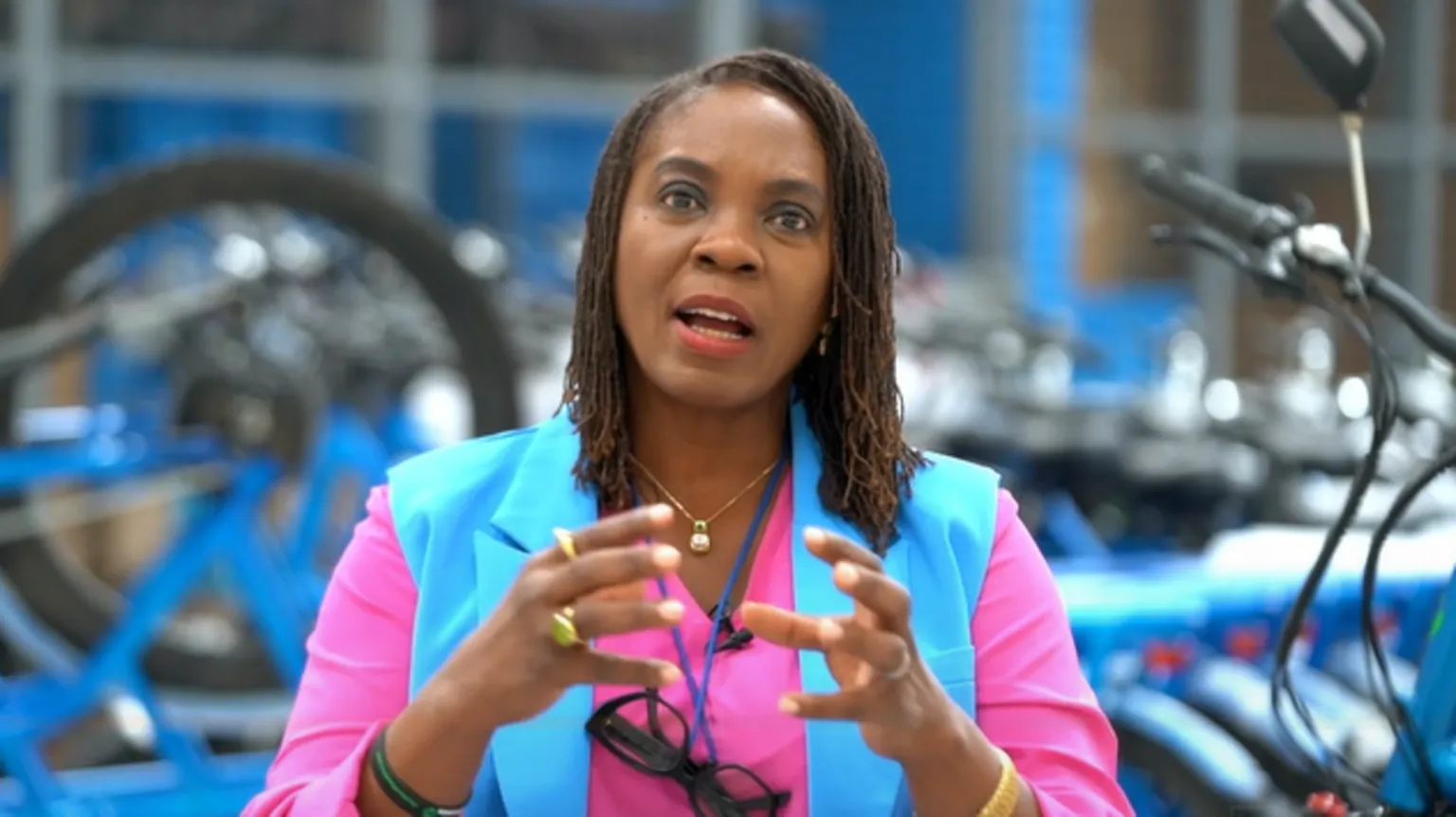 A black woman dressed in a bright pink and blue top, holding up her hands as if gesturing while speaking, with rows of bicycles out of focus behind the woman