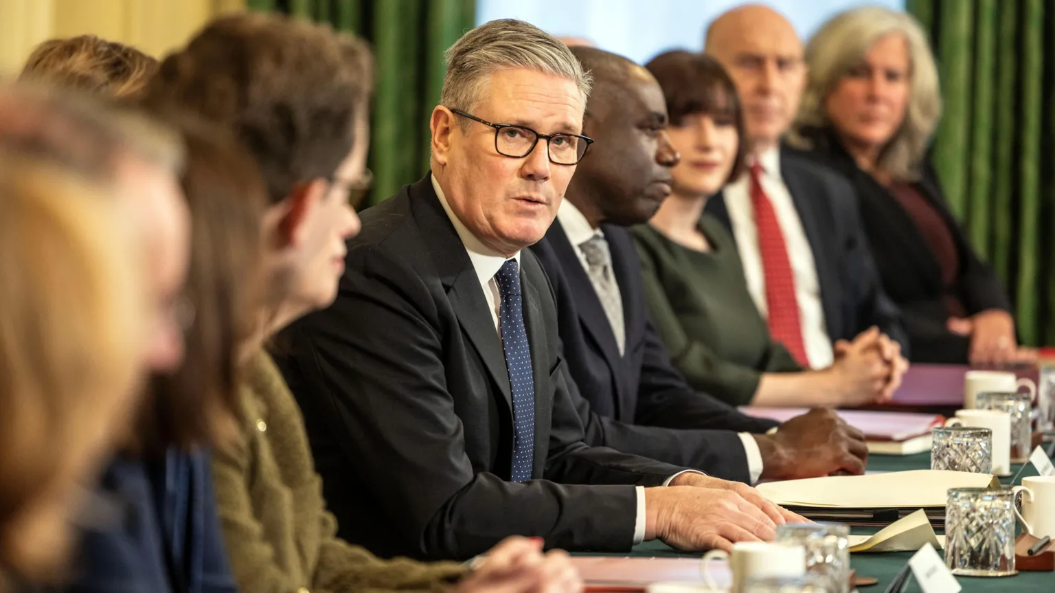 Sir Keir Starmer addresses members of the cabinet. He is looking towards the camera and is wearing glasses and a dark suit and tie with a white shirt. Some members of the cabinet are looking away from the camera and some are not in focus. In front of them are papers, drinks glasses and mugs.