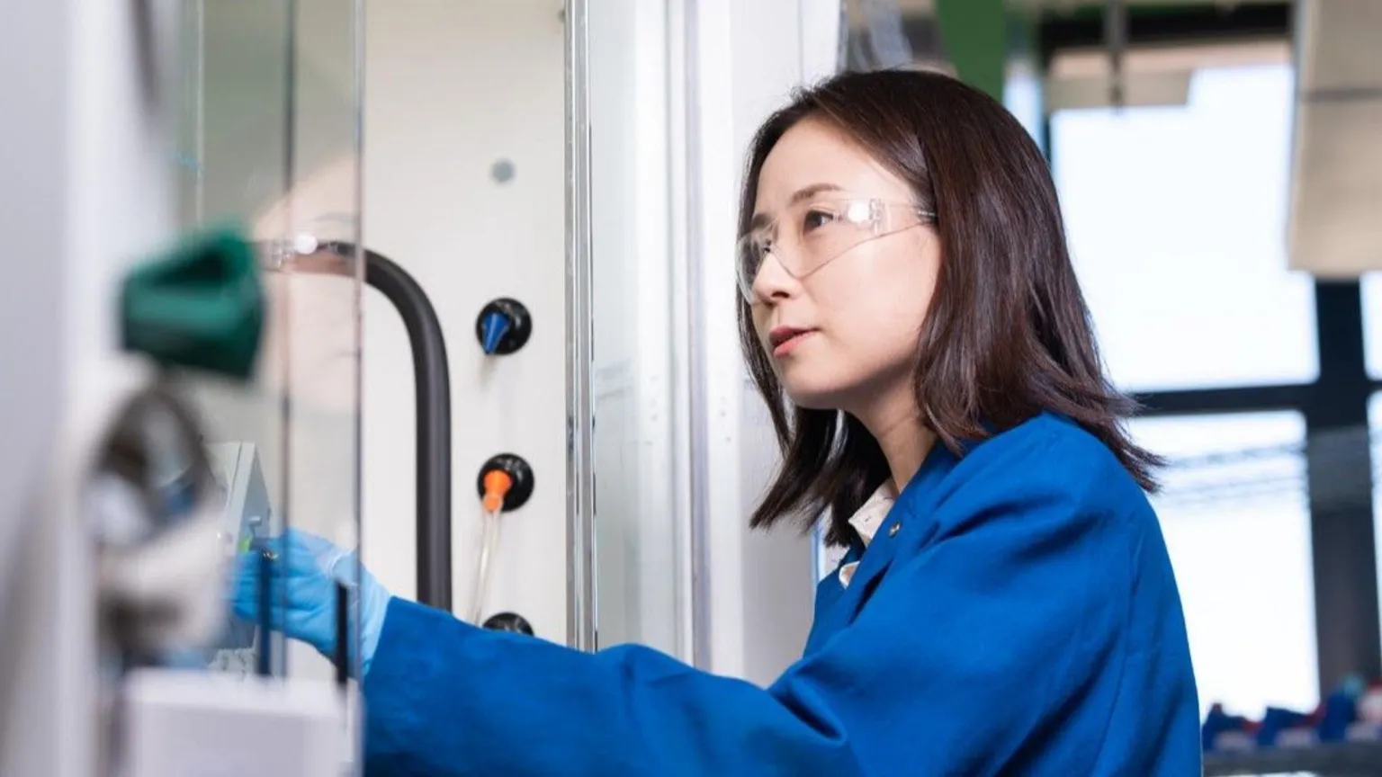 Jeff Liang, UCSB Professor Grace Han in blue overalls looks towards a piece of scientific equipment.