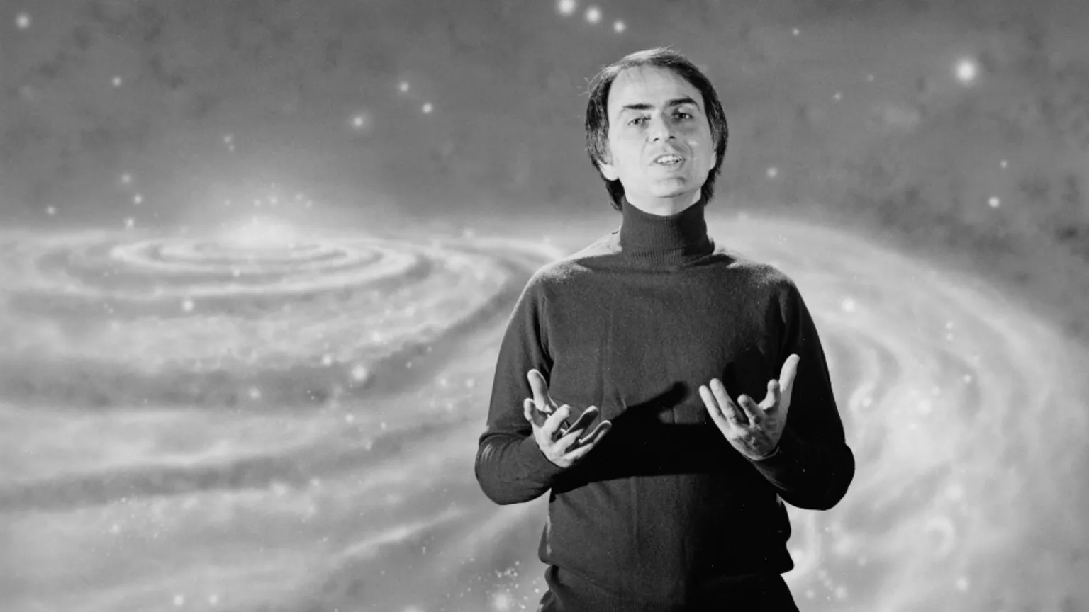 Getty Images Black and white image of Carl Sagan, who has combed over dark hair and is wearing a turtle neck jumper. Behind him is a graphic of the universe.