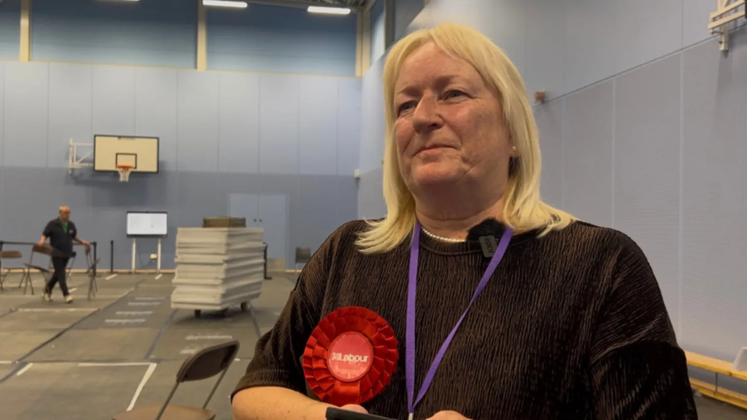 A woman wearing a red Labour badge stood inside a leisure centre looking to the side of the camera