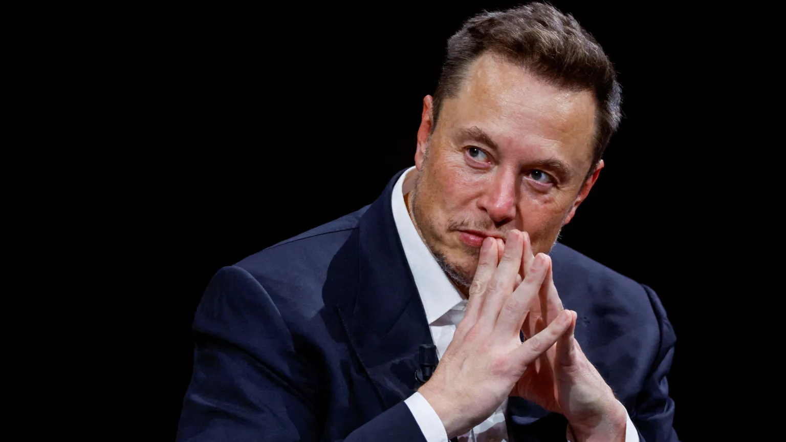 Reuters Elon Musk on a stage with his hands tented in front of his mouth as he listens to someone next to him.