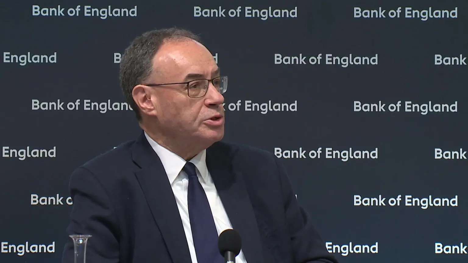 Bank of England governor Andrew Bailey speaking at a news conference. He is wearing a dark blue suit, white shirt and dark blue tie, and sitting in front of a screen with 'Bank of England' written on it