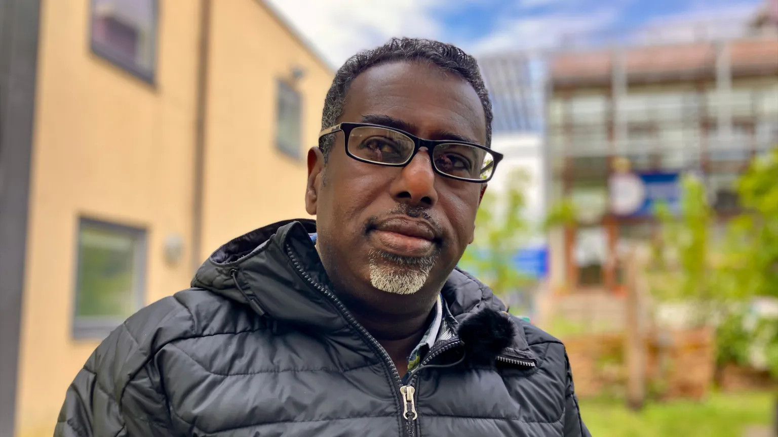 Yassin Mohamud wearing black glasses and a black puffer jacket, looking directly at the camera. The background is blurred, but there are buildings and greenery behind him.