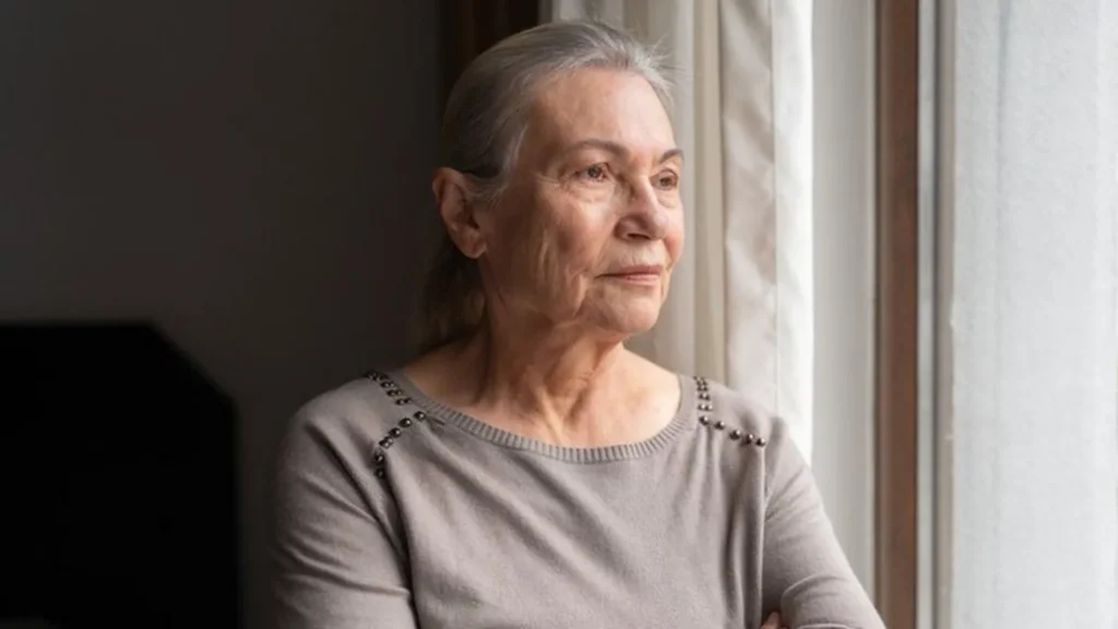 Getty Images An older woman with long silver hair looks out a bedroom window. She wears a pale grey top with silver beads on the shoulders.