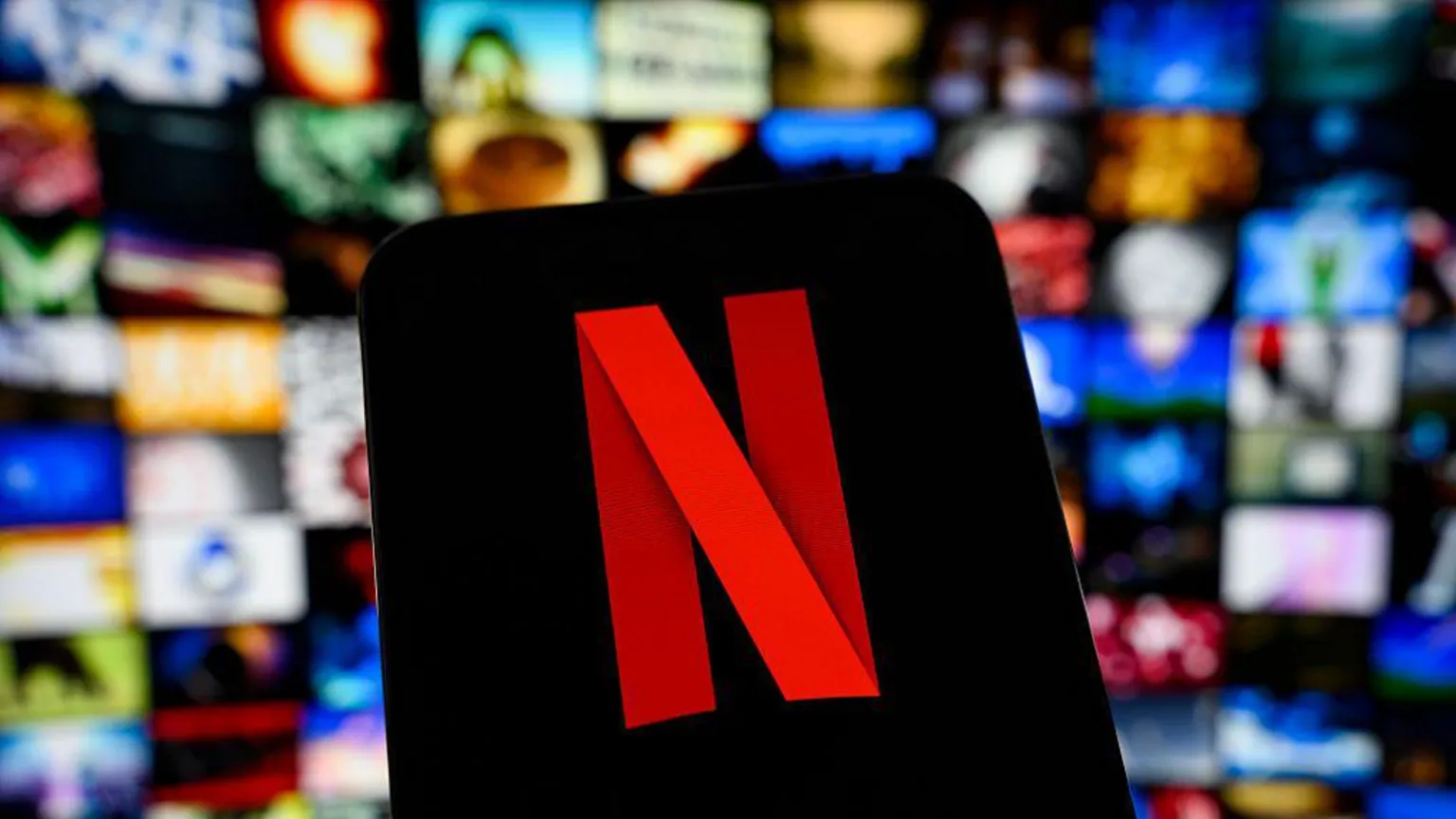 Netflix logo shown on a smartphone screen, with the platform and its rows of film and TV shows shown blurred in the background behind it.