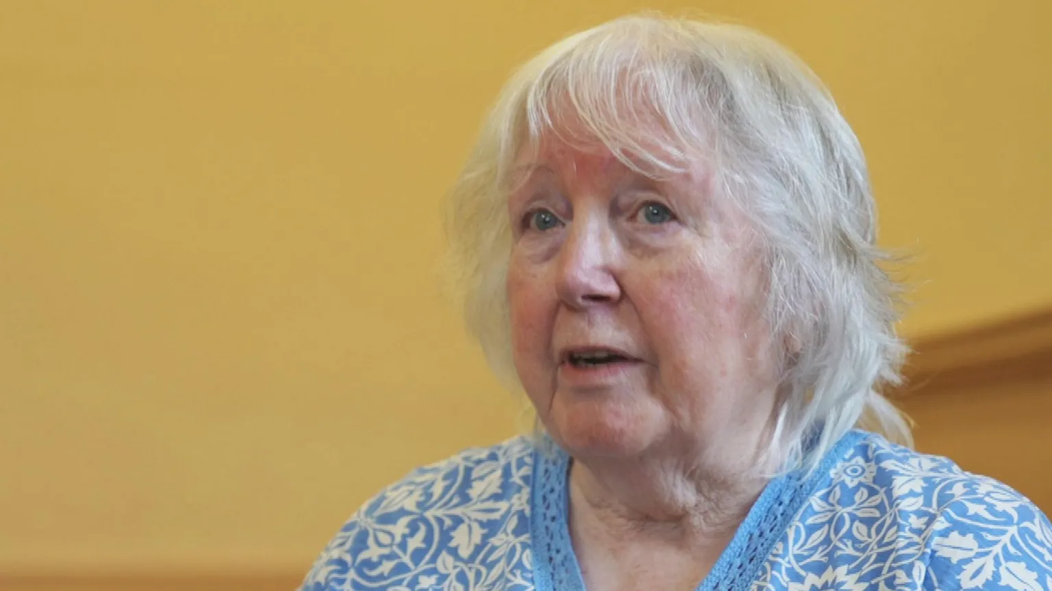 Margaret Gent, a woman with shoulder-length grey hair, wearing a blue top with a white pattern. There are yellow walls in the background.