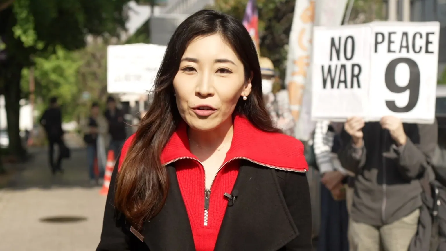 BBC correspondent Kurumi Mori wearing a red top and black coat standing on the street next to a person holding a sign that reads "no war, peace 9"