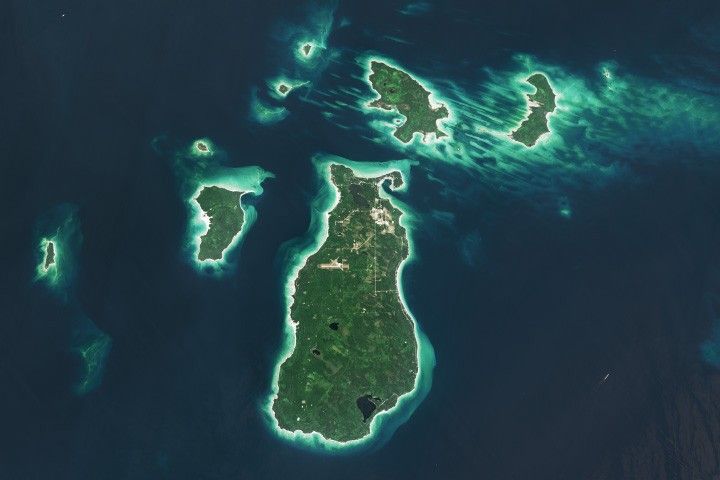 A satellite image shows several islands in Lake Michigan. The largest one, Beaver Island, is in the center. The islands are mostly green and vegetated, with bright sandy areas on their perimeters. Shallow waters near the land appear turquoise, and deeper waters are dark blue.