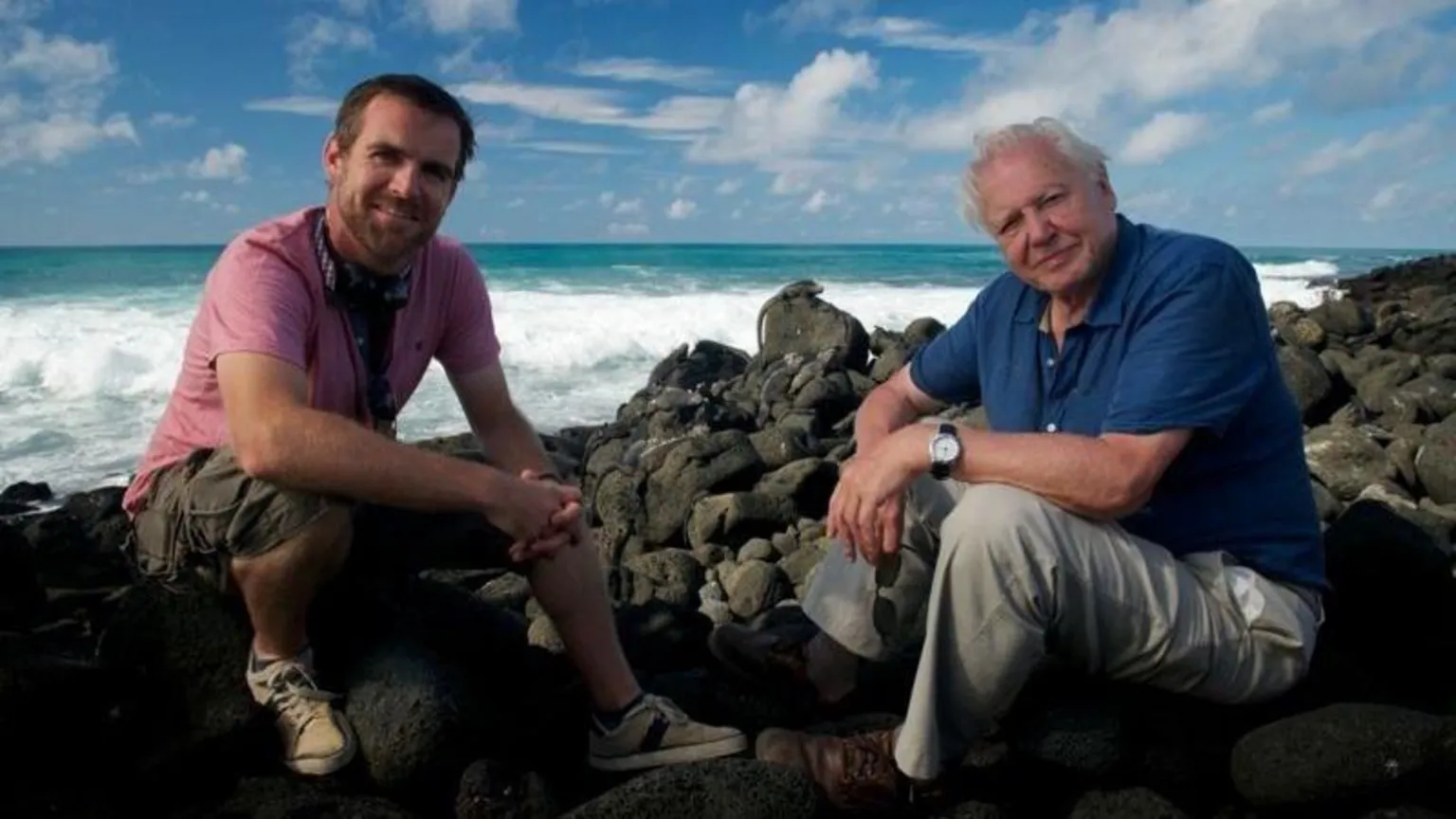 Martin Williams and David Attenborough sat next to on another on rocks next to the ocean.