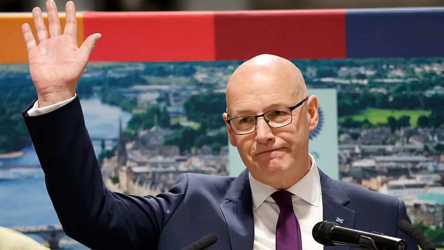 Getty Images John Swinney, in blue suit, white shirt and purple tie, smiles and waves as he stands on a podium with a photo of Perth behind him.